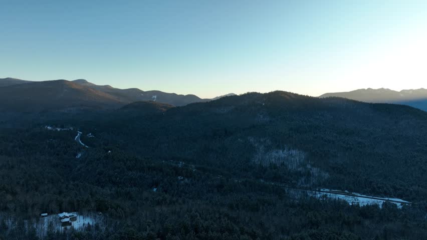 A top-down drone shot over a snow-covered forest in the Adirondack Mountains, revealing a narrow road and a white modern house nestled among frost-covered evergreen trees.