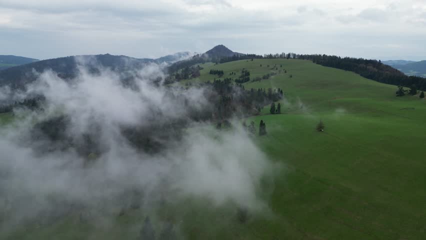 Aerial drone flight over green hills of the Pieniny Mountains near Szczawnica after rainfall. Low clouds and fog drift above meadows and forests, creating calm, cinematic landscape toward Wysoka Peak.