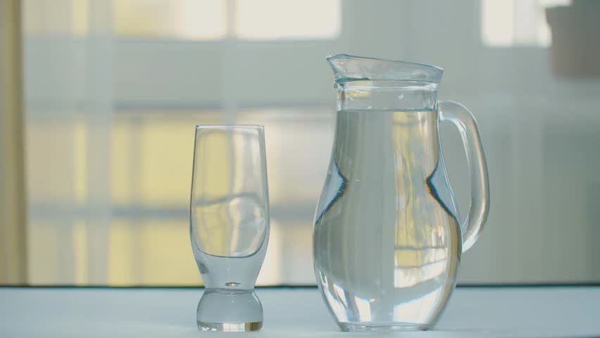 Woman’s hand pouring fresh water from a jug into a glass on a table, symbolizing healthy hydration, wellness, diet, and lifestyle habits.