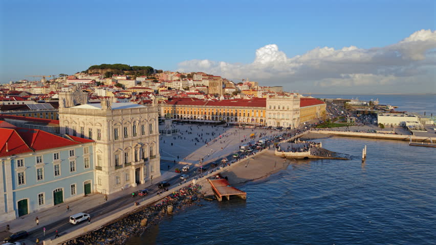 Drone footage shows Praca do Comercio from above with riverside promenade, moving crowds and iconic Lisbon buildings, highlighting scale, symmetry and urban space