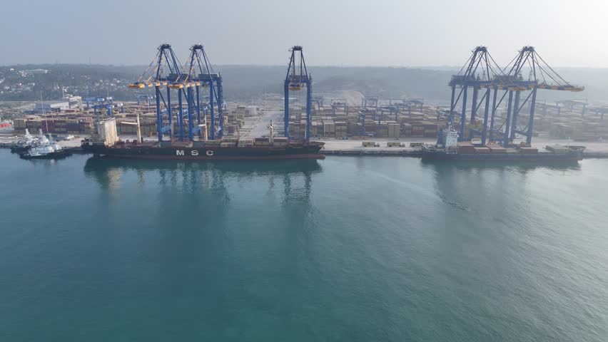 Aerial drone footage of a large container port near Kovalam in Kerala, India. Cargo ships docked at the terminal with container cranes, stacked shipping containers and maritime logistics infrastructure. Industrial harbor scene showing global trade, transportation and shipping operations.