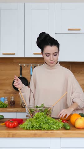 Attractive young woman with a hair bun adding fresh cherry tomatoes to a glass bowl and mixing a colorful vegetable salad with wooden spoons on a countertop in a modern bright kitchen