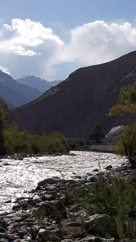 The video captures a serene river flowing through Khunjerab National Park in the Hunza region of Pakistan. Crystal-clear mountain water rushes over rocky beds, surrounded by rugged brown hills, green bushes, and distant snowy peaks under a bright blue sky with fluffy clouds. Natural water sounds create a peaceful, relaxing atmosphere in this high-altitude Karakoram landscape.