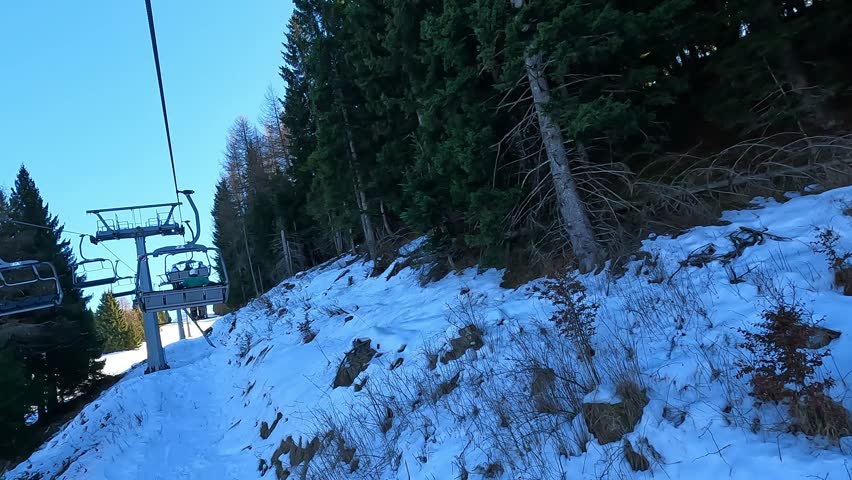 Side view from a chairlift passing close to tall pine trees on a steep snowy mountainside, capturing the immersive experience of riding a ski lift through a winter forest on a sunny day.