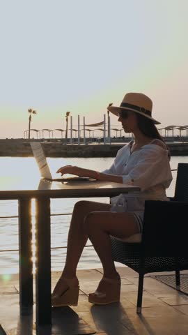 Young woman in summer clothes, sunglasses and sun hat, uses laptop, sitting alone at table on promenade by the sea, at sunset or sunrise. freelancer, remote work. summer vacation at sea