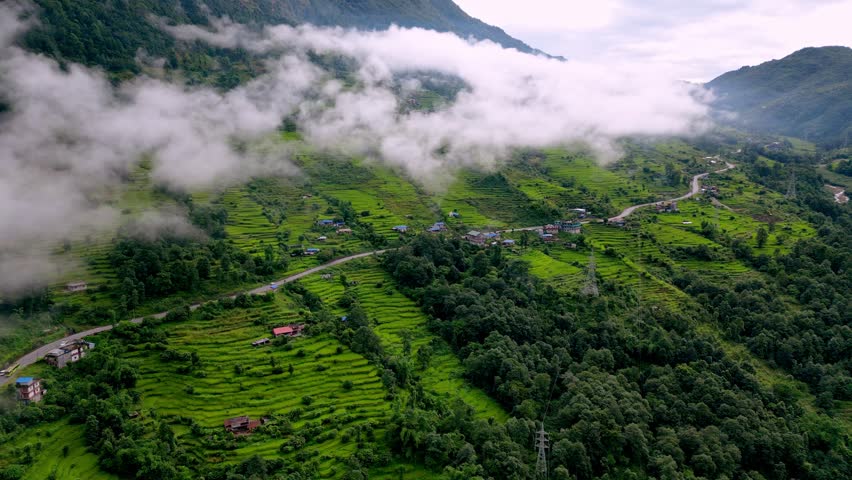 Rice terraces and a winding road in the rural mountains of Nepal, surrounded by mist and low clouds. Cinematic aerial drone footage showing village life and lush green landscapes.