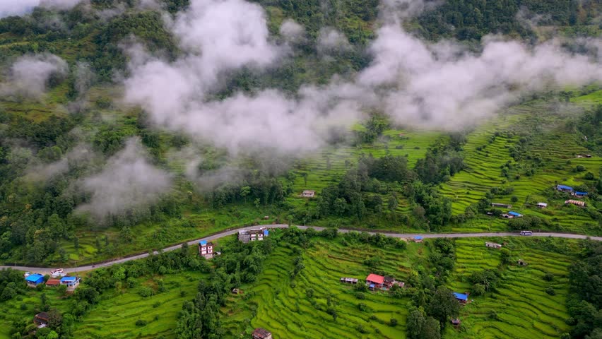 Rice terraces and a winding road in the rural mountains of Nepal, surrounded by mist and low clouds. Cinematic aerial drone footage showing village life and lush green landscapes.