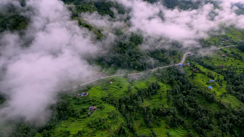 Rice terraces and a winding road in the rural mountains of Nepal, surrounded by mist and low clouds. Cinematic aerial drone footage showing village life and lush green landscapes.