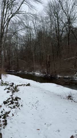 Point of view while walking down a small snow-covered hill in a home backyard with Cherry Creek flowing through a forest in the winter in Stroudsburg, Pennsylvania in the Pocono Mountains