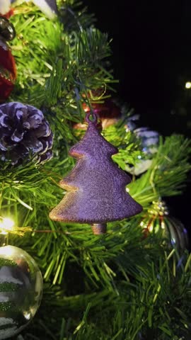 Close-up shot of a Christmas tree ornament in the shape of a tree hanging on a beautifully decorated Christmas tree with glowing fairy lights. Vertical screen