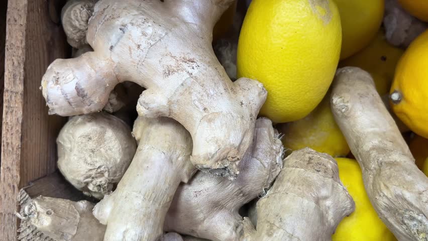 Horizontal video of fresh ginger roots and lemon in one wooden crate, followed by garlic bulbs and onions with natural peel remnants in another crate. Slow left-to-right camera movement captures textures and realistic market presentation. Concept of cooking ingredients, healthy food, and grocery shopping.