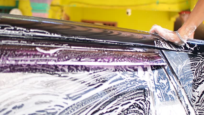 Close-up of a person's hand washing a black car with soap suds, scrubbing the roof during a car wash service.
