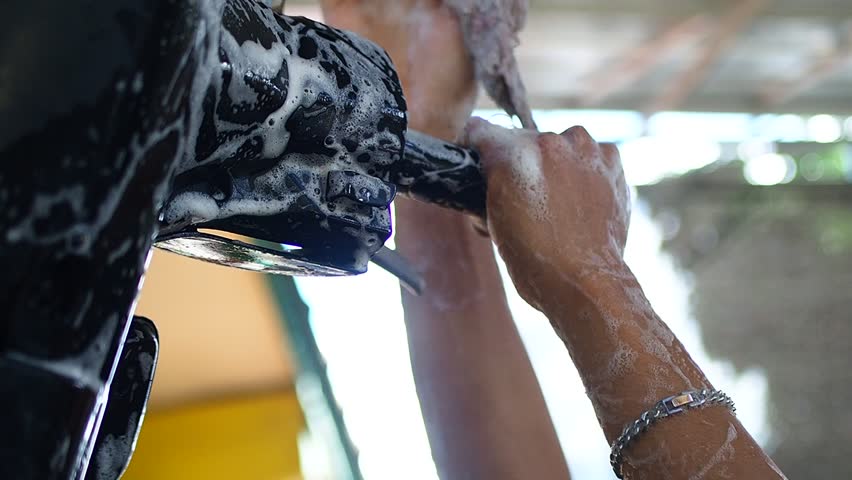 Close-up of a person's hand washing a black motorcycle handle covered in white soap suds