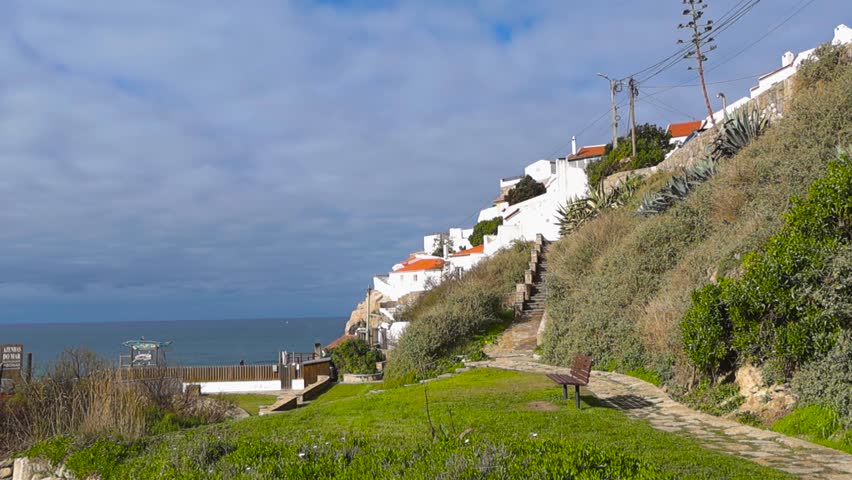 Smooth panoramic pan reveals Azenhas do Mar with white cliffside homes, red rooftops, grassy park paths, and the Atlantic Ocean stretching beneath a bright, lightly clouded sky.