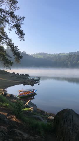 A peaceful vertical shot of a calm lake in the early morning. The scene features thick white mist rising from the water, with colorful wooden boats docked along the rocky shore. A pine tree branch frames the view, with lush green hills in the distance under a clear blue sky.