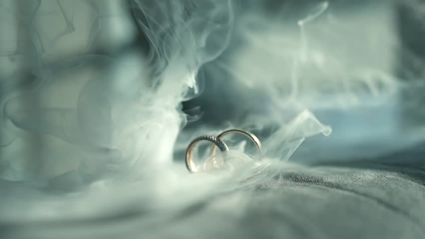 Static detail close-up shot of two gold wedding rings resting on soft white lace or tulle fabric with soft natural light and smoke effect subtle shadow creating romantic wedding ring detail.