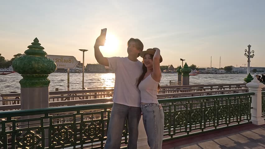 Smiling couple takes smartphone selfie at riverside promenade during golden hour sunset, handheld camera movement