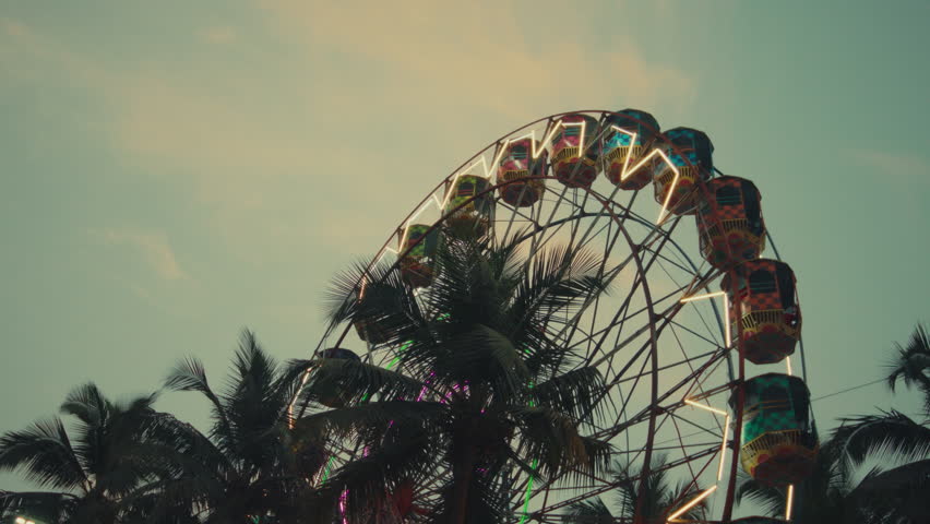 A cinematic shot of a Ferris wheel with neon lights spinning behind palm trees at dusk. Captures the nostalgic charm and vibrant energy of a seaside amusement park.