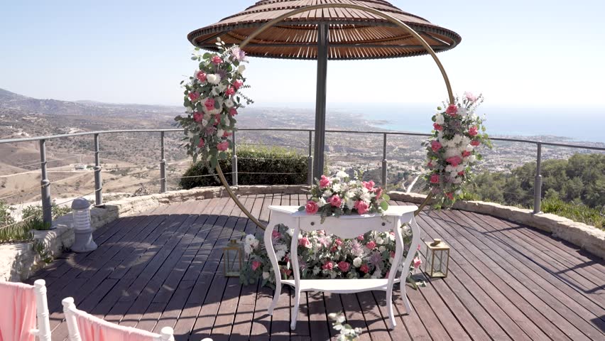 A wedding setup takes place on a terrace with flowers and a circular arch. The coast and hills are visible in the background. Chairs are arranged for guests.