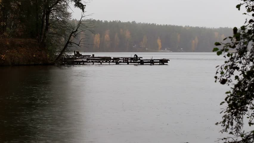 Outdoor scene of tranquil lake during rain. Dilapidated wooden pier stretches into water from tree-lined bank. Forest of autumn trees visible on far shore under grey sky.
