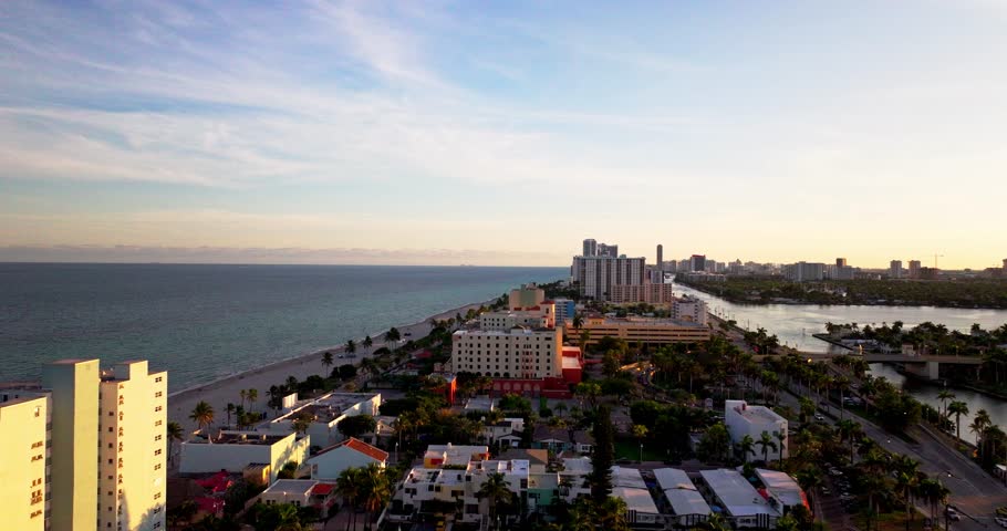 Smooth aerial flyover of Hallandale Beach, Florida during golden hour, showcasing beachfront residential buildings, palm-lined streets, inland waterways, and the Atlantic Ocean coastline.