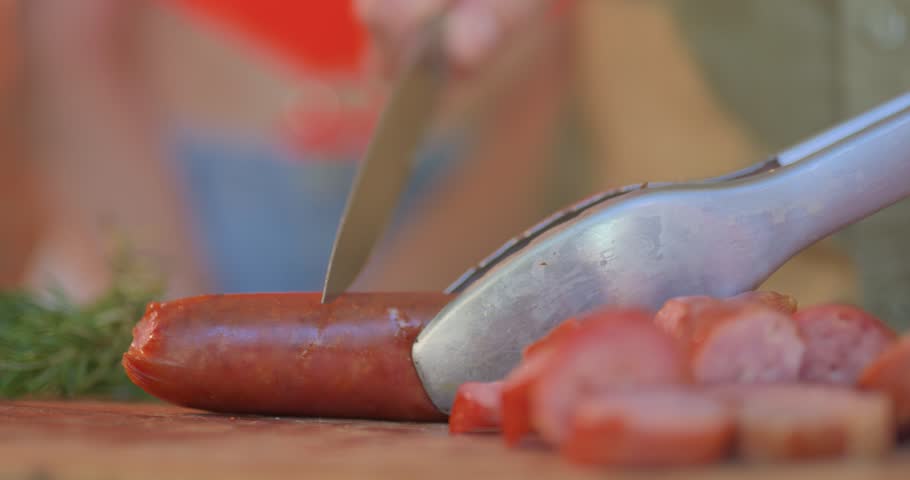 Close up hands slicing grilled chorizo on wooden board as friends pick and share bites with toothpicks, summer backyard barbecue party and good vibes.