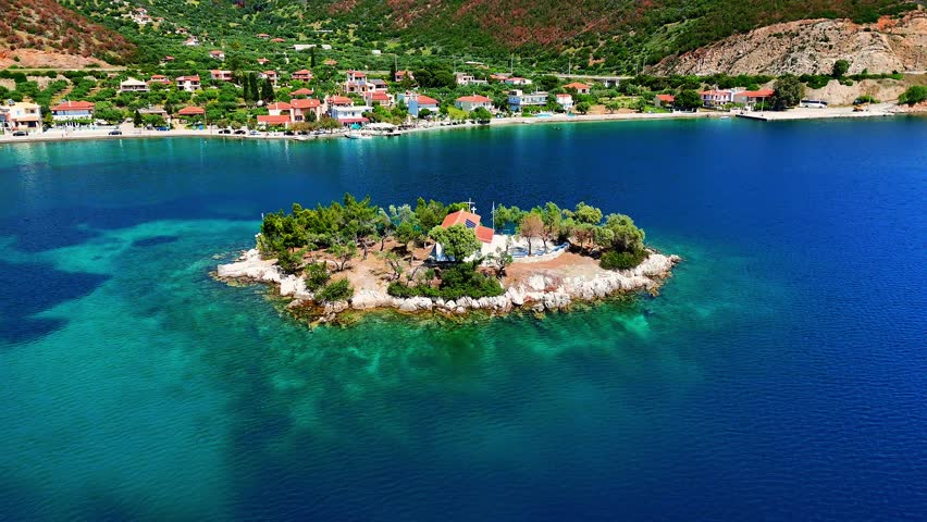 Aerial view of the small islet of Agios Nikolaos on the Delphi Riviera, Greece, surrounded by clear turquoise water, rugged coastline and green hills under a bright summer sky.