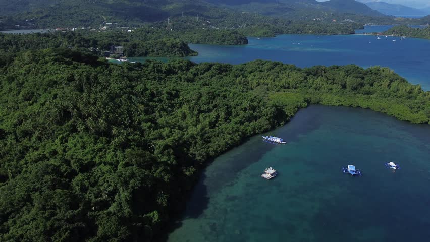 Aerial drone footage of a tropical bay in Puerto Galera Philippines, with lush green islands and forested mountains in the background, calm blue water and anchored boats in a serene natural landscape.