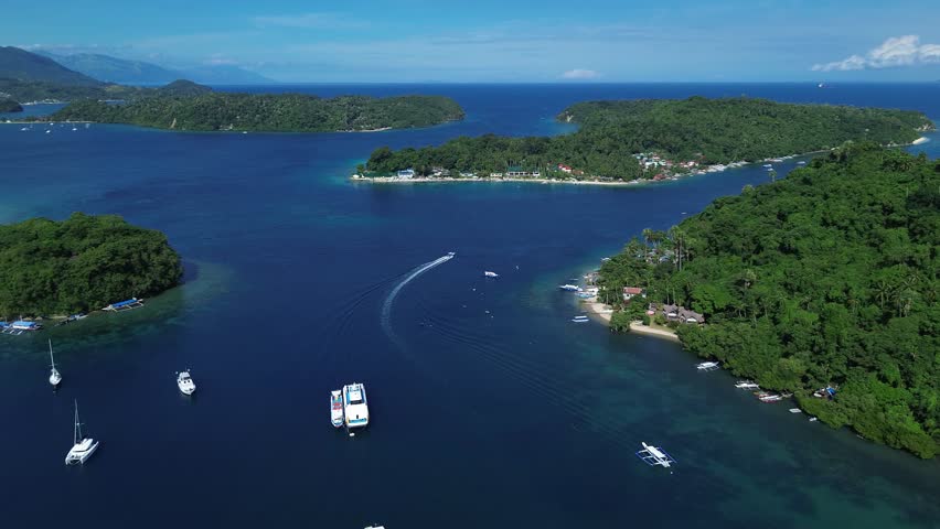 Aerial drone footage of a tropical bay in Puerto Galera Philippines, with lush green islands and forested mountains in the background, calm blue water and anchored boats in a serene natural landscape.