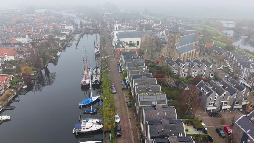 Aerial view of foggy Dutch canal town gabled brick homes, docked boats, and tall church tower form a tranquil, symmetrical waterfront scene softened by mist.