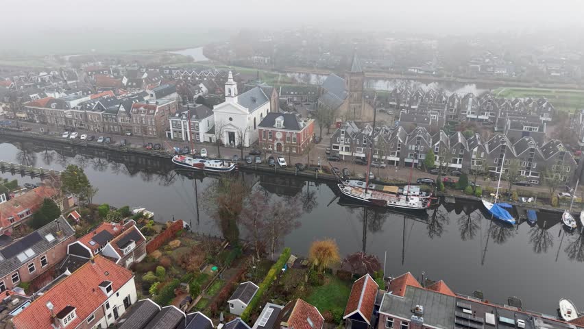 Aerial view of foggy Dutch canal town gabled brick homes, docked boats, and tall church tower form a tranquil, symmetrical waterfront scene softened by mist.