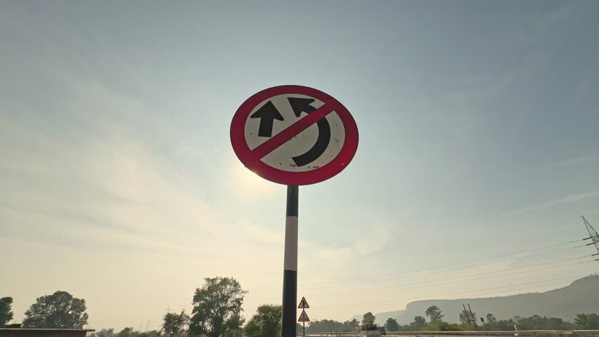 Static shot of a no U-turn road sign mounted on a pole, framed against clear sky with distant trees and highway elements in soft daylight.