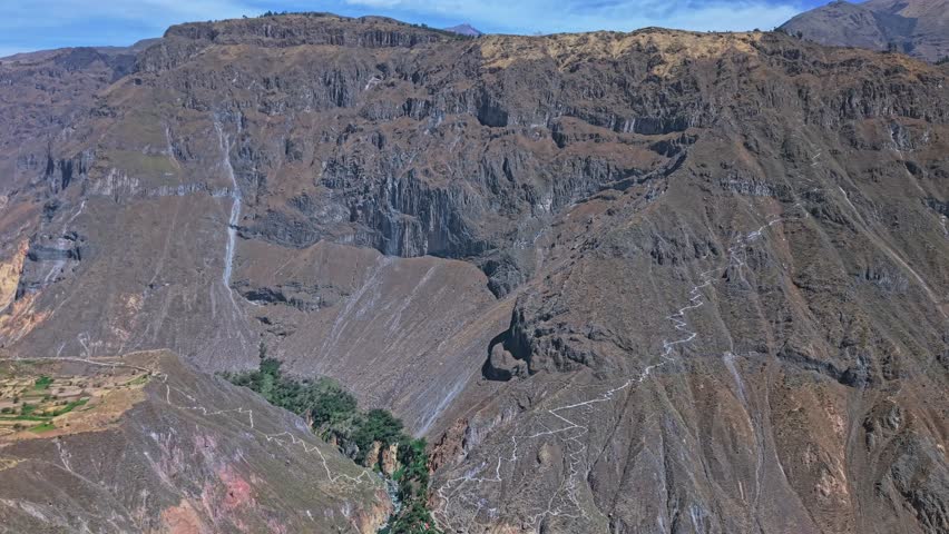 Drone pans left to right over the Colca Canyon entrance trail descending from the canyon top toward the oasis enormous sheer cliffs and the full canyon height emphasise scale adventure rugged terrain.