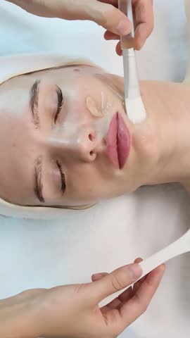Cosmetologist applies clear gel to cheek of young beautiful caucasian woman using white brush during professional facial skincare procedure in modern clinic calm beauty concept clean aesthetic care