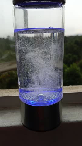 This is a hydrogen water generator bottle in operation. The device uses electrolysis to infuse molecular hydrogen into the water. The swirling motion of the microbubbles at the start of the video shows the rapid release of gas from the internal electrodes. The bottle has a glass transparent body and a metallic base hydrogen, visible as a dense cloud of fine white bubbles rising from the base through the water. 