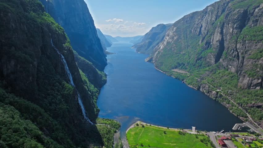High angle drone view of Geirangerfjord surrounded by towering cliffs in Norway. Cinematic footage shows dramatic fjord depth, green slopes and remote village far below.
