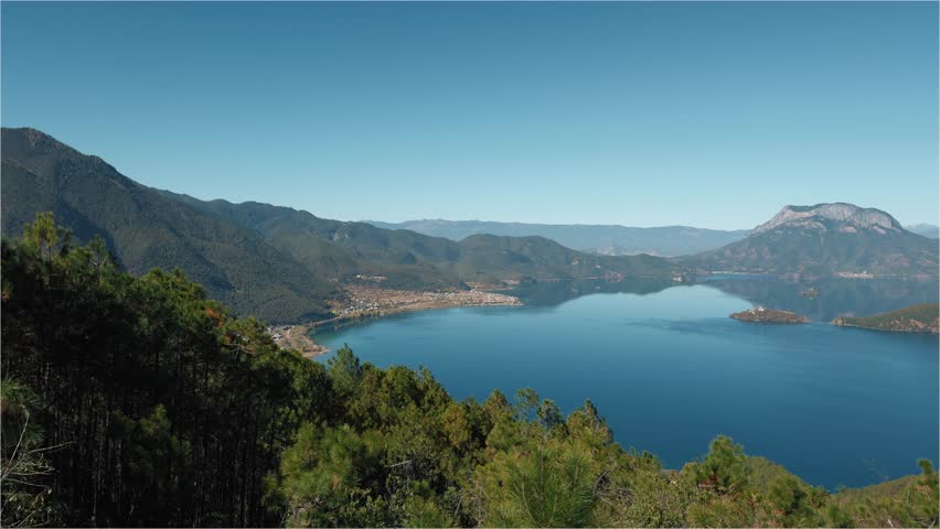 A wide panoramic view of Lugu Lake reveals deep blue waters encircled by forested mountains under clear skies in Yunnan, China.