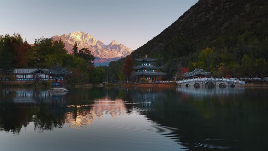 The first sunlight paints Jade Dragon Snow Mountain in gold as Black Dragon Pool reflects traditional pavilions, arched bridges, and calm water in a serene dawn scene.