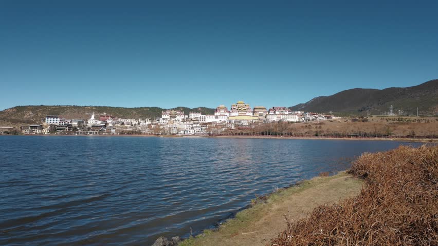 The grand Songzanlin Monastery rises above a calm lakeside in Shangri-La, showcasing iconic Tibetan architecture set against clear skies and mountain scenery.