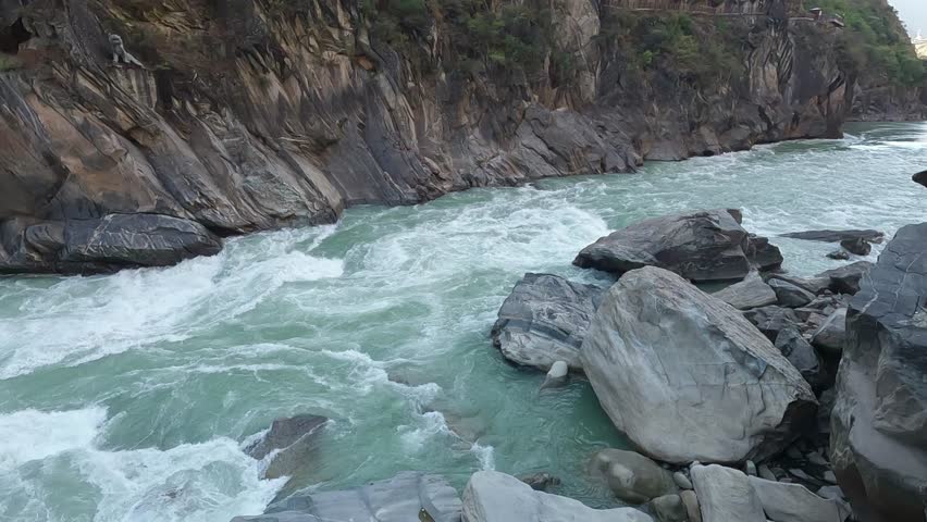 Powerful turquoise rapids surge between massive rocks in Tiger Leaping Gorge, showcasing the raw force of the Yangtze River cutting through a steep mountain canyon.