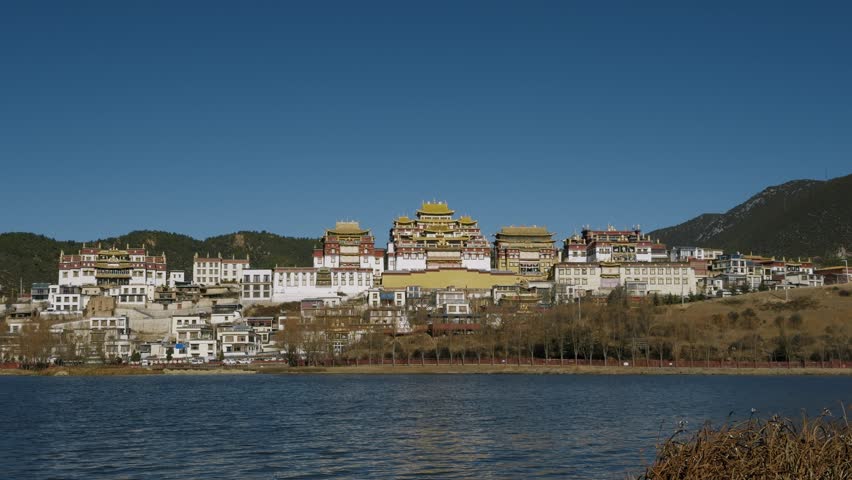The grand Songzanlin Monastery rises above a calm lakeside in Shangri-La, showcasing iconic Tibetan architecture set against clear skies and mountain scenery.