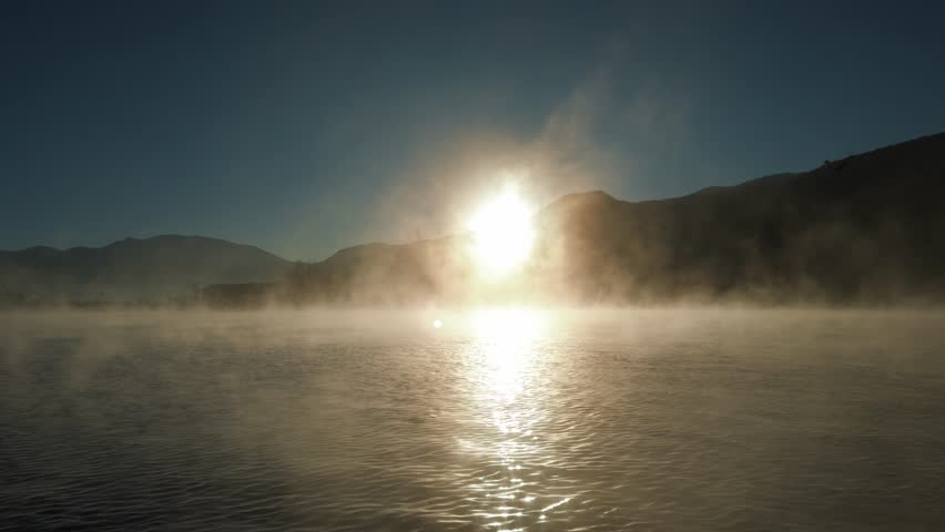 Soft morning mist drifts across Lugu Lake as the rising sun illuminates calm water and distant mountains, creating a tranquil dawn atmosphere.