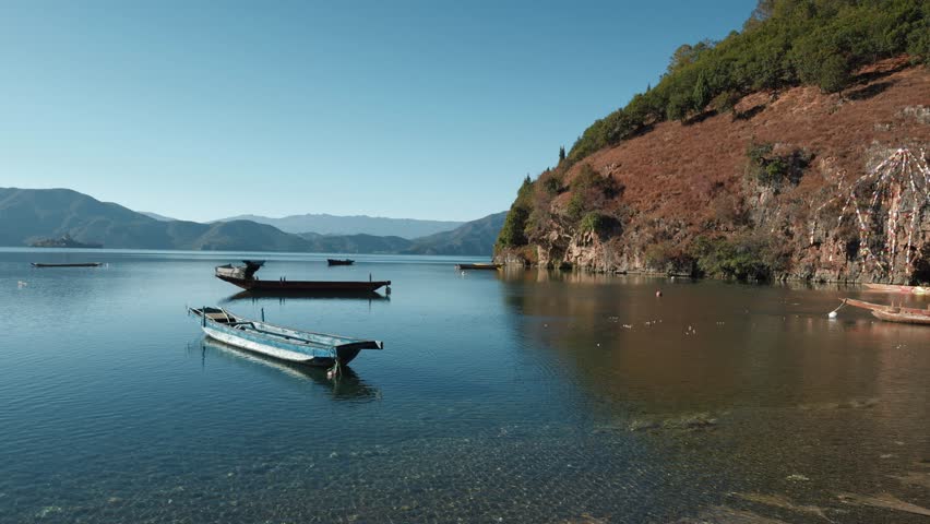 Small wooden boats rest on the calm waters of a clear mountain lake, framed by forested hills and distant peaks under a bright blue sky.