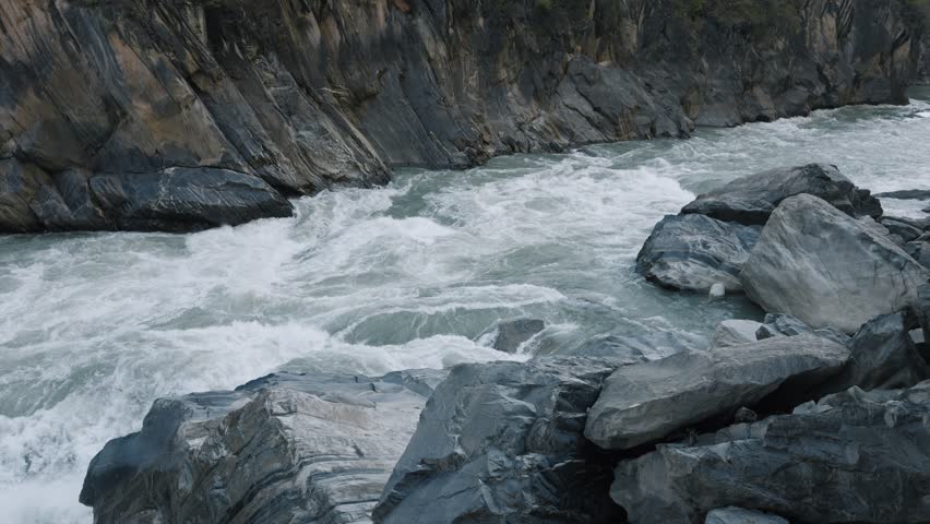 Powerful turquoise rapids surge between massive rocks in Tiger Leaping Gorge, showcasing the raw force of the Yangtze River cutting through a steep mountain canyon.