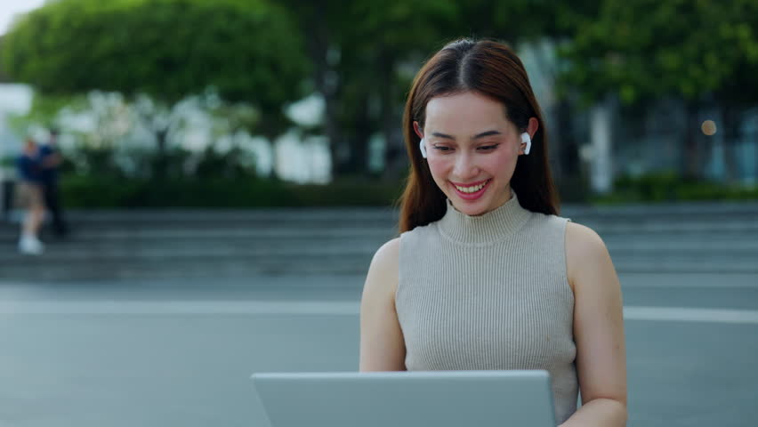 Portrait of Asian woman with earphones joins online lesson via laptop in city park. Friendly Korean lady waves hand to classmates on video call by computer sitting in downtown