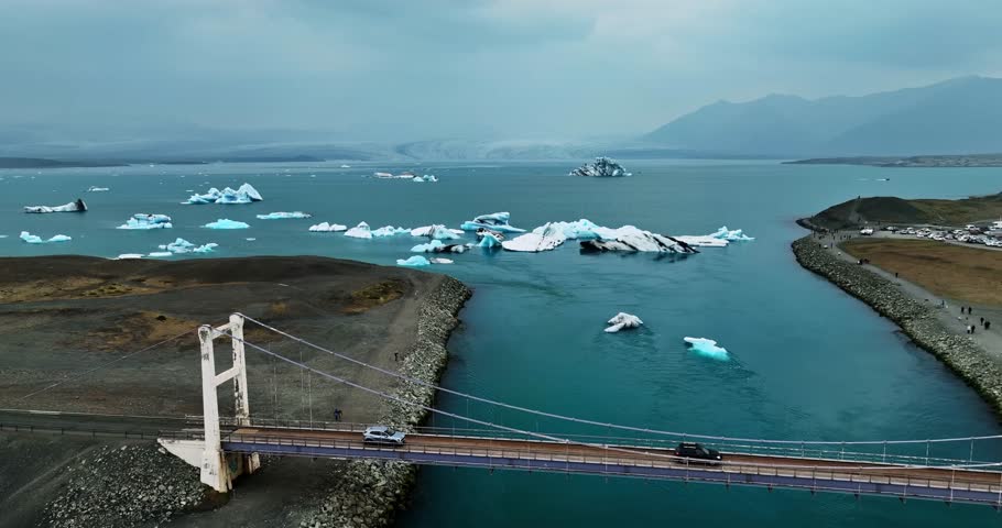 Drone aerial over Jokulsarlon lagoon outlet in Iceland, suspension bridge and causeway framing drifting blue icebergs in calm glacial water under cloudy skies.