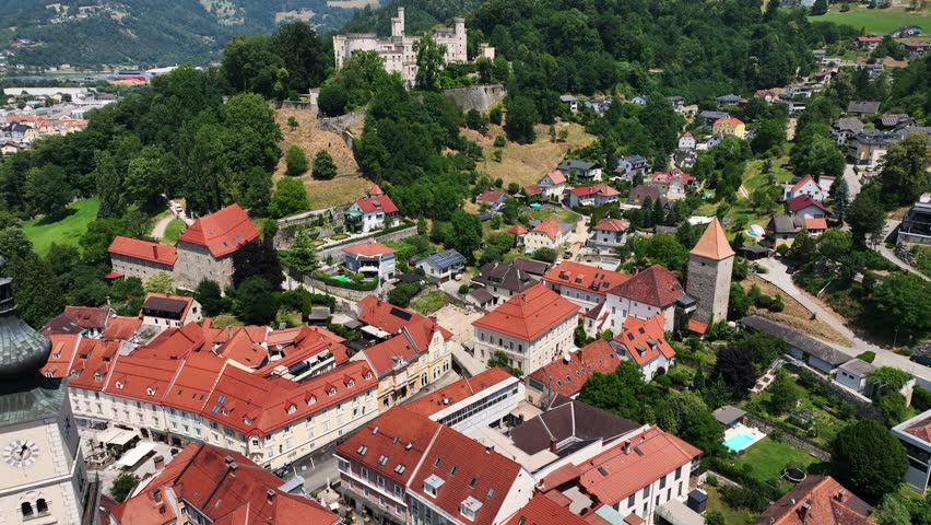 Aerial summer footage of Wolfsberg showing town center, church towers, green hills, layered valleys, warm light, smooth drone flight, atmospheric European small-town scenery