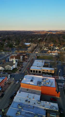An aerial view captures the charming town center of Plainfield, Illinois, at sunset, showcasing a bustling main street flanked by historic buildings and a prominent church steeple.