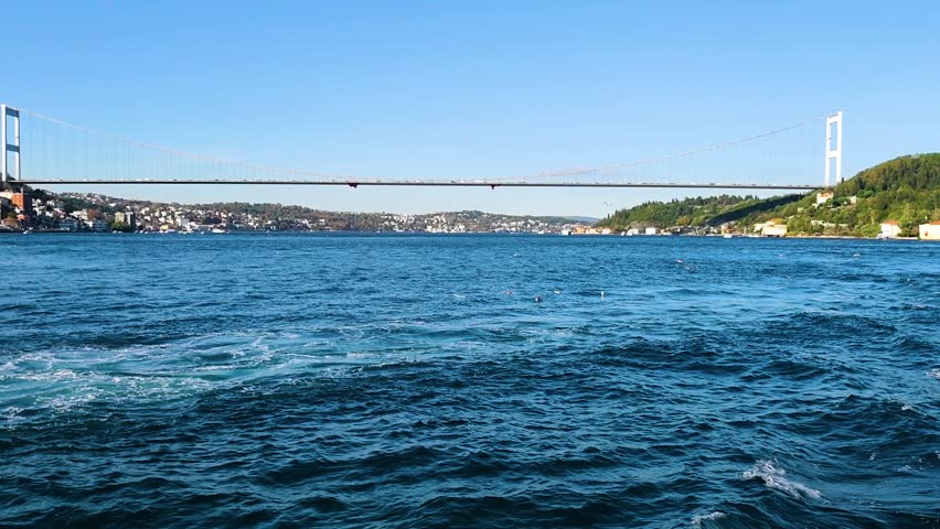 Captivating high-quality footage of the Fatih Sultan Mehmet Bridge (Second Bosphorus Bridge) in Istanbul, featuring a lively flock of seagulls soaring over the sparkling blue waters. The video is shot from a moving boat, capturing the energetic white foam and turquoise ripples of the Bosphorus Strait in the foreground. The suspension bridge stands as a majestic landmark in the background, perfectly connecting the European and Asian continents under a clear, brilliant blue sky.

The scene beautifully illustrates the vibrant harmony between nature and urban engineering. This clip is an ideal choice for travel documentaries, tourism advertisements for Turkey, maritime transportation themes, and promotional videos showcasing the scenic beauty of Istanbul. The natural, bright daylight highlights the architectural details of the bridge and the fluid motion of the birds, providing a cinematic and uplifting atmosphere. Whether you are creating content for a global city guide, an environmental piece on marine life, or a professional background for broadcast media, this authentic footage captures the true spirit and energy of one of the world