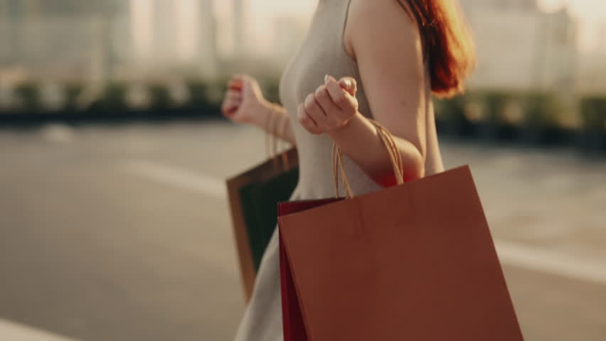 Crop view of woman with shopping bags hanging on arms walks across urban square slow motion. Young female carries purchases in paper packages at holiday sale in town district